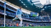 A Manchester City player taking a corner kick at Etihad Stadium during a match. The player is wearing a light blue kit and the stadium is filled with a large crowd of spectators.