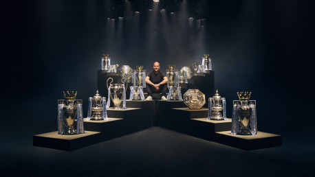 A person is sitting surrounded by various football trophies, including Premier League, FA Cup, and UEFA Champions League trophies.