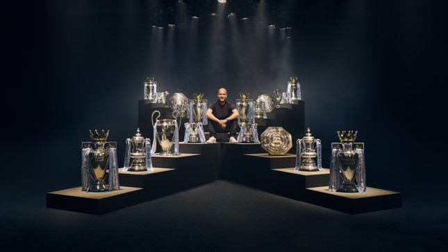 A person is sitting surrounded by various football trophies, including Premier League, FA Cup, and UEFA Champions League trophies.