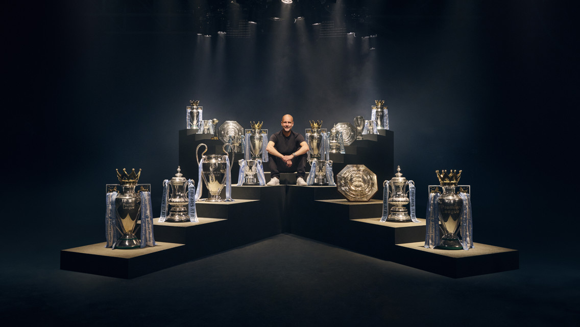 A person is sitting surrounded by various football trophies, including Premier League, FA Cup, and UEFA Champions League trophies.