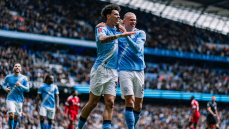 Two soccer players in light blue jerseys celebrating a goal in a stadium with a crowd.