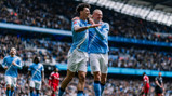 Two soccer players in light blue jerseys celebrating a goal in a stadium with a crowd.