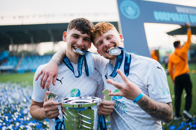 Two football players wearing white Puma jerseys with medals around their necks, holding a trophy at a Corporate Cup event. Two football players wearing white Puma jerseys with medals around their necks, holding a trophy at a Corporate Cup event.
