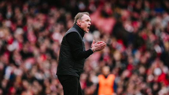 A coach wearing black is clapping on the sidelines during a match with a blurred crowd in the background.