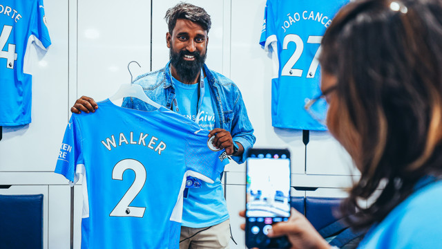 A person is holding a Manchester City jersey with the name 'WALKER' and number 2 in a locker room, while another person takes a photo using a smartphone.