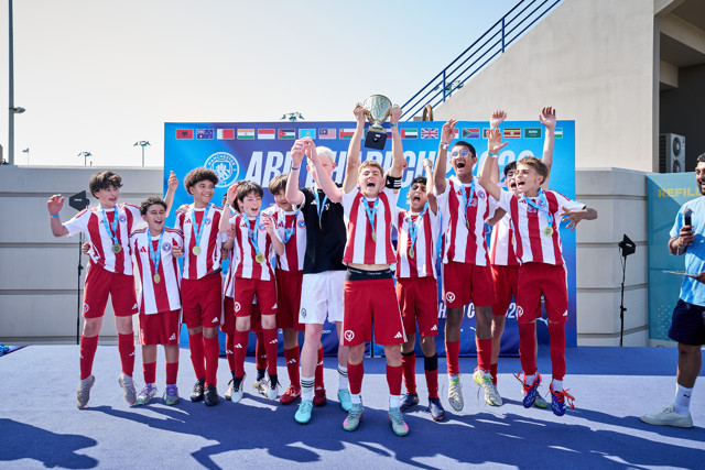 A group of young soccer players in red and white uniforms celebrating with a trophy on a stage. Flags from various countries are displayed in the background.