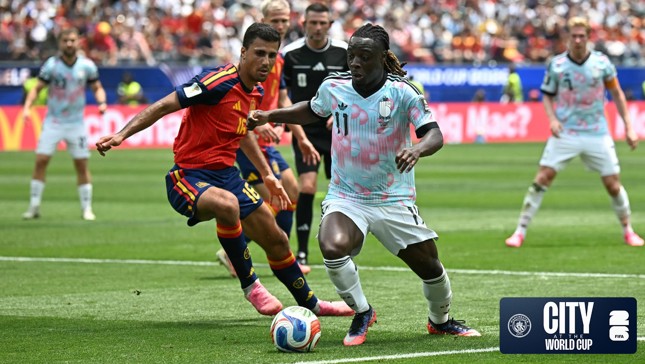 Two soccer players from opposing teams, one in a red and blue Spain jersey and the other in a light patterned jersey, compete for the ball during a match. The field is green and there are spectators in the background.