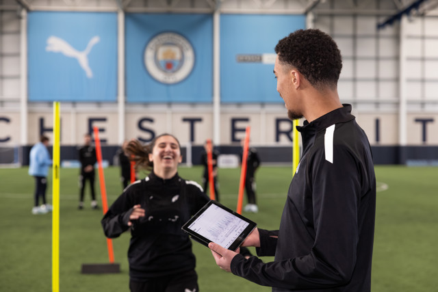 Individuals participating in a training session indoors at a soccer facility, utilizing equipment and technology for performance analysis. The background features the Manchester City logo.