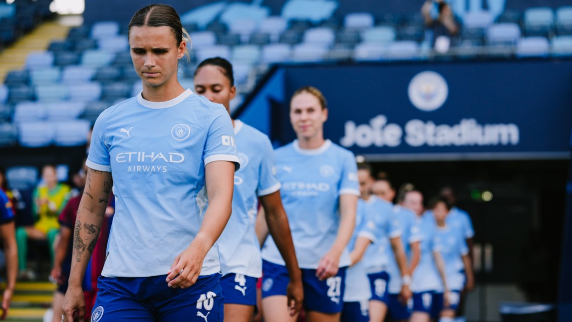 Manchester City women's team walking out at Joie Stadium with players in focus wearing light blue jerseys.