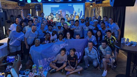 A group of Manchester City supporters wearing team jerseys pose together holding a large 'Official Supporters Club' flag with Manchester City logos, celebrating a gathering indoors.