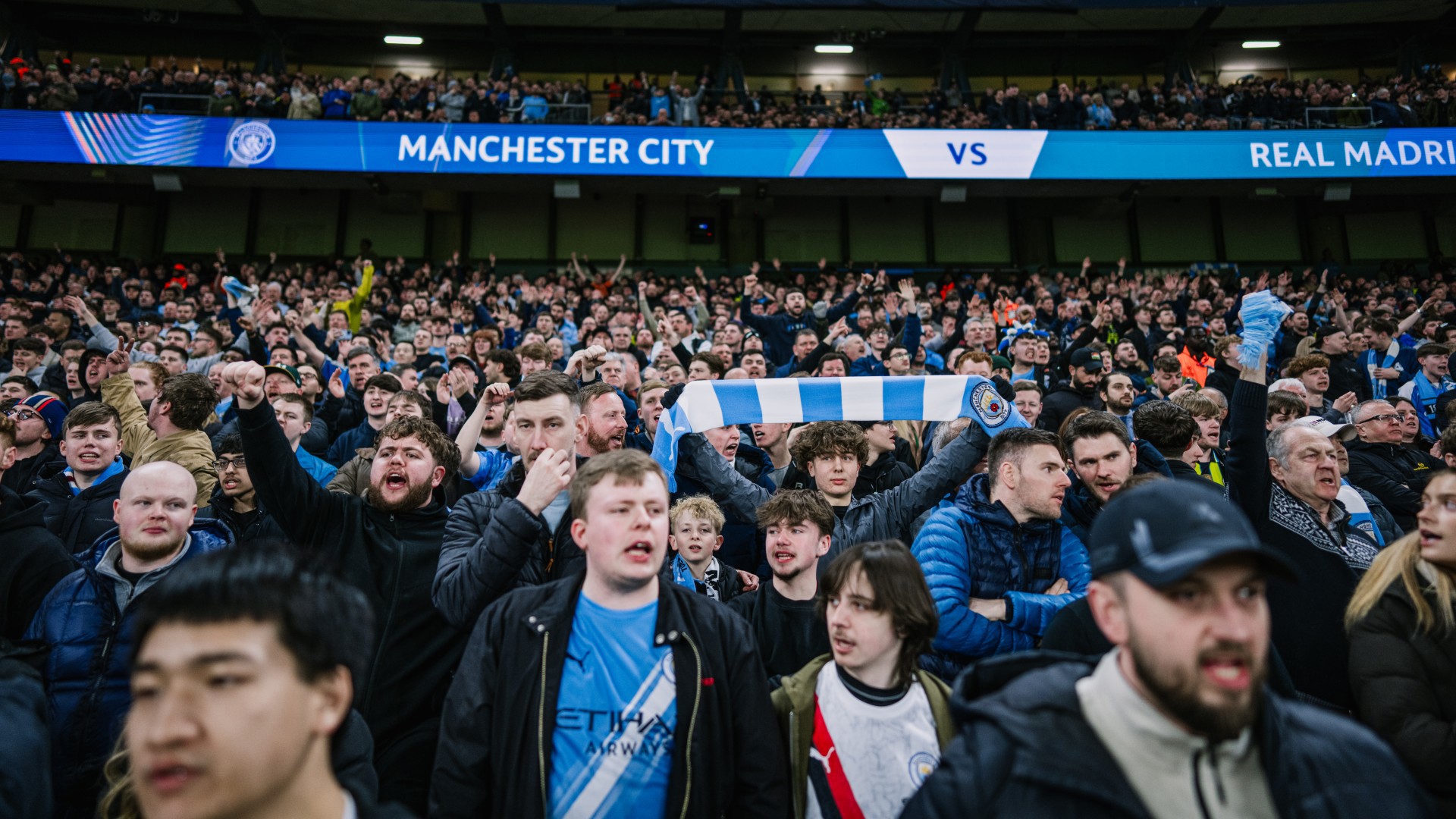 A crowd of Manchester City fans cheering at a football match against Real Madrid. A fan holds a Manchester City scarf. The stadium is filled with enthusiastic supporters.