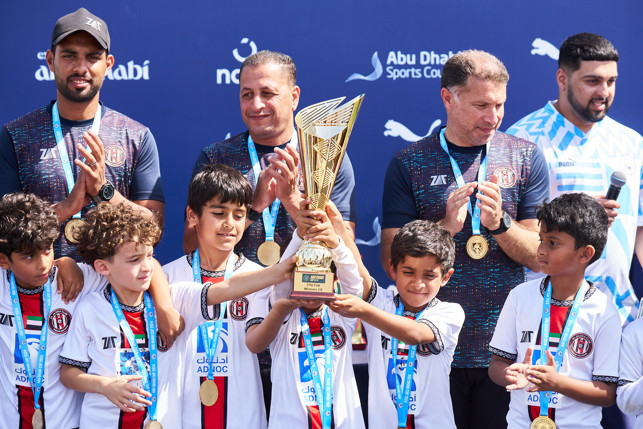 Young children celebrating with a trophy in hand alongside their coaches against a backdrop labeled Abu Dhabi Sports Council.