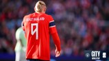 A footballer wearing a red jersey with 'BRAUT HAALAND' and number 9 on the back stands on a crowded field. Manchester City and FIFA World Cup logos are visible.