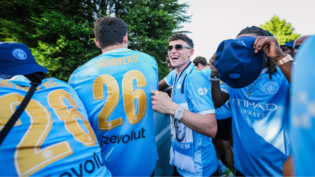 Group of people wearing Manchester City jerseys with 'WINNERS' and number 26 on the back. A man adjusts his hat, and others wear scarves. They are outdoors with trees in the background.