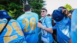 Group of people wearing Manchester City jerseys with 'WINNERS' and number 26 on the back. A man adjusts his hat, and others wear scarves. They are outdoors with trees in the background.