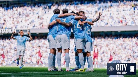 A group of Manchester City players in blue jerseys are celebrating a goal in a stadium filled with spectators. They are huddled together with arms around each other. The background features blurred cheering fans.