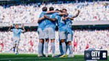 A group of Manchester City players in blue jerseys are celebrating a goal in a stadium filled with spectators. They are huddled together with arms around each other. The background features blurred cheering fans.