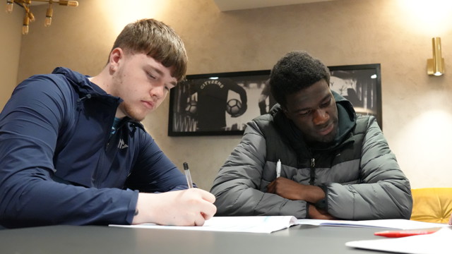 Two people engaged in paperwork discussion, one writing notes, seated in a room with Manchester City memorabilia on the wall.