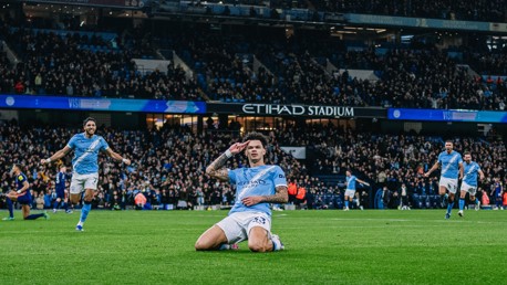 A Manchester City player celebrates after scoring a goal at Etihad Stadium, with other teammates running behind. The crowd is visible in the background.