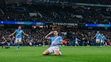 A Manchester City player celebrates after scoring a goal at Etihad Stadium, with other teammates running behind. The crowd is visible in the background.
