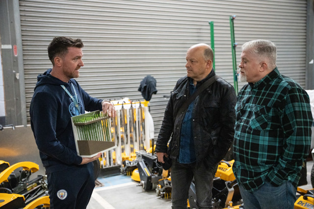 Three men engage in a discussion inside a warehouse. One man holds a display model with grass-like features. Manchester City logo visible on clothing.