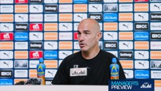 A person speaking at a press conference with a backdrop featuring Manchester City sponsors and logos. Lucozade bottles are visible on the table.