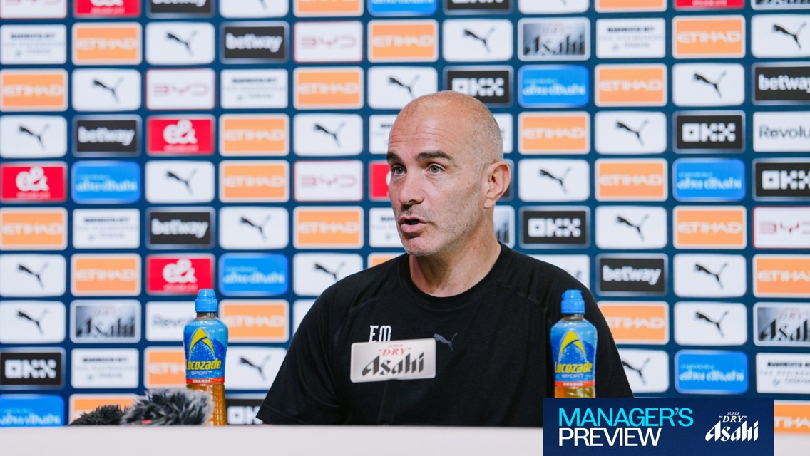 A person speaking at a press conference with a backdrop featuring Manchester City sponsors and logos. Lucozade bottles are visible on the table.