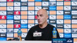 A person speaking at a press conference with a backdrop featuring Manchester City sponsors and logos. Lucozade bottles are visible on the table.