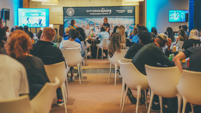 A seminar event scene with attendees seated in white chairs, facing a stage where a panel is speaking. The backdrop displays logos including Man City Electric Blue Logo and Etihad.