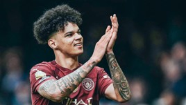 A football player claps wearing a maroon Manchester City jersey, adorned with tattoos on his arms.