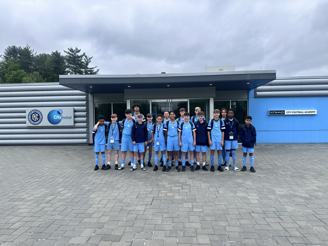 A group of young athletes wearing light blue football kits stand in front of a building with City Football and New York City Football Club logos. The location appears to be the Etihad City Football Academy.