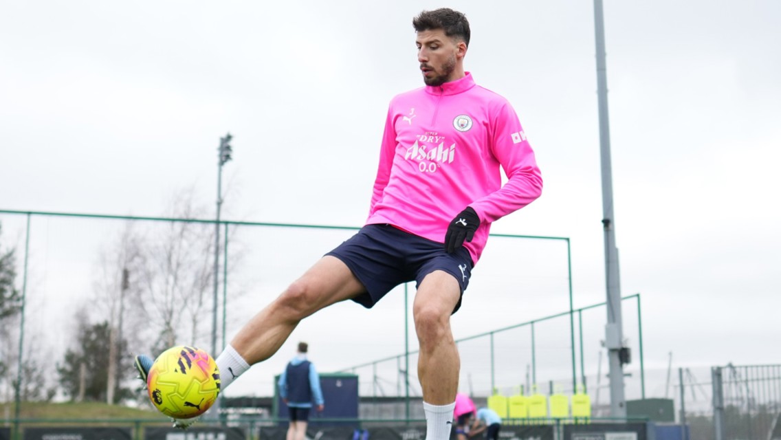 A soccer player in a bright pink Manchester City training top performs a skill move with a ball on a training field.