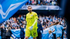 A goalkeeper wearing a Manchester City jersey with number 25 walks on the pitch at a football match, surrounded by fans and blue banners.