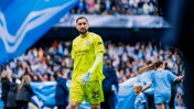 A goalkeeper wearing a Manchester City jersey with number 25 walks on the pitch at a football match, surrounded by fans and blue banners.