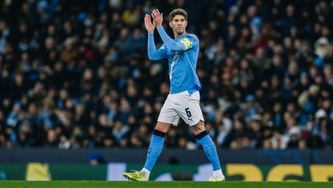 A football player in a light blue kit with the number 5, wearing a captain's armband, claps towards the crowd during a match.