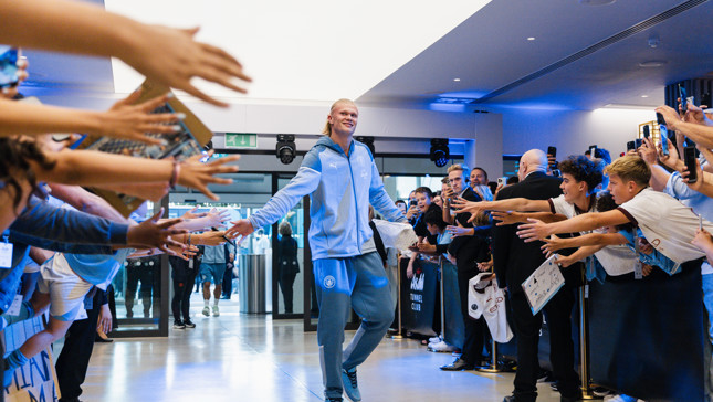 A player in a Manchester City tracksuit is walking through a tunnel, high-fiving fans on both sides.