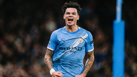 A football player in a Manchester City kit, with visible tattoos, celebrating during a match.
