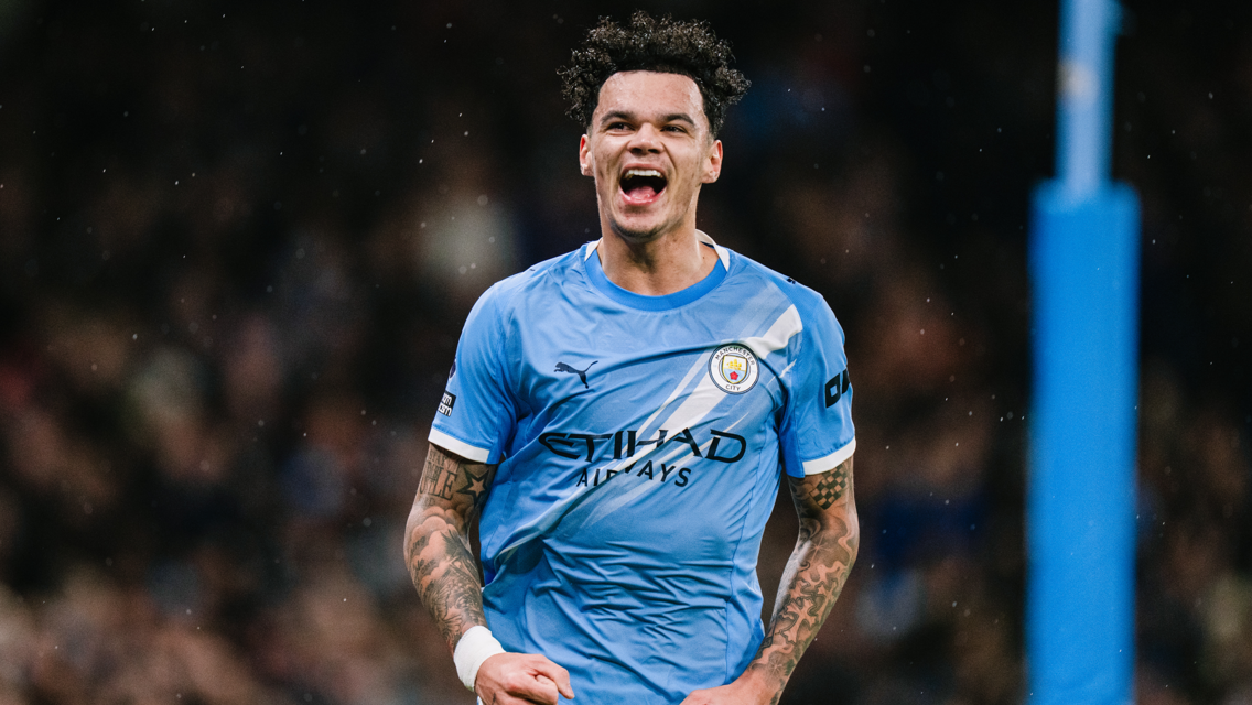 A football player in a Manchester City kit, with visible tattoos, celebrating during a match.