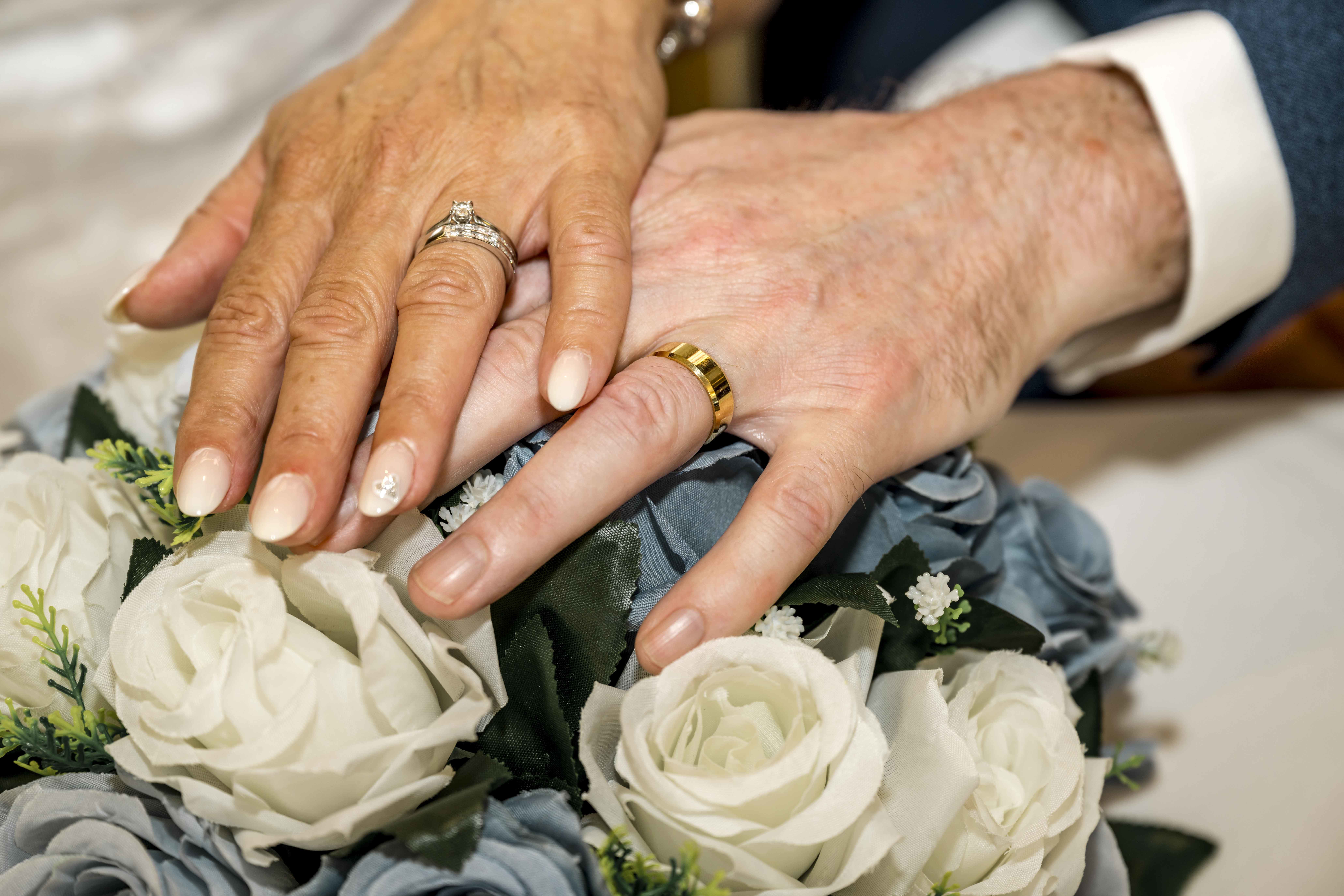 A close-up image showing two hands with wedding rings resting on a bouquet of blue and white flowers.