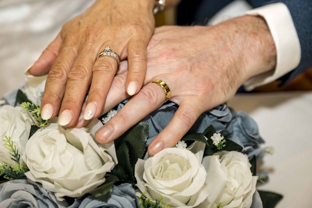 A close-up image showing two hands with wedding rings resting on a bouquet of blue and white flowers.