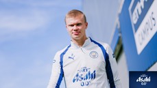 A person in a Manchester City training outfit with the Asahi 0.0% logo, standing outdoors against a blue sky.