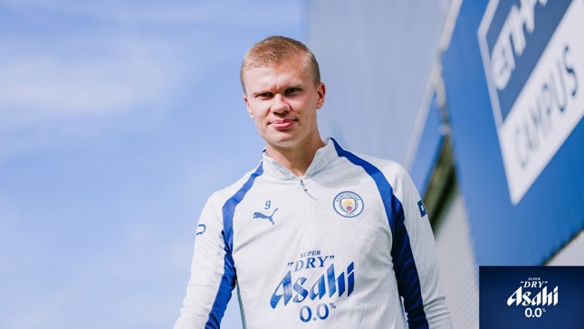 A person in a Manchester City training outfit with the Asahi 0.0% logo, standing outdoors against a blue sky.