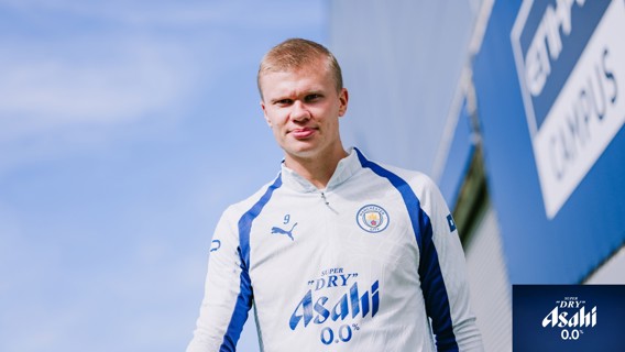 A person in a Manchester City training outfit with the Asahi 0.0% logo, standing outdoors against a blue sky.