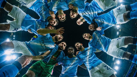 A team huddle of Manchester City football players wearing their light blue jerseys, viewed from below with stadium lights in the background.