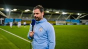 A person in a Manchester City tracksuit holding a microphone on a football field with blurred face.