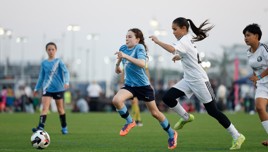 Youth soccer players competing on a field. A player in a blue jersey runs towards the ball, chased by opponents wearing white jerseys.