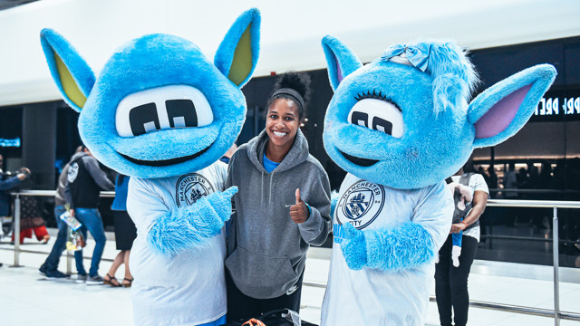 Two people dressed as Man City mascots Moonbeam and Moonchester pose for a photo with a blurred individual in the center. The mascots are wearing shirts with the Manchester City logo.