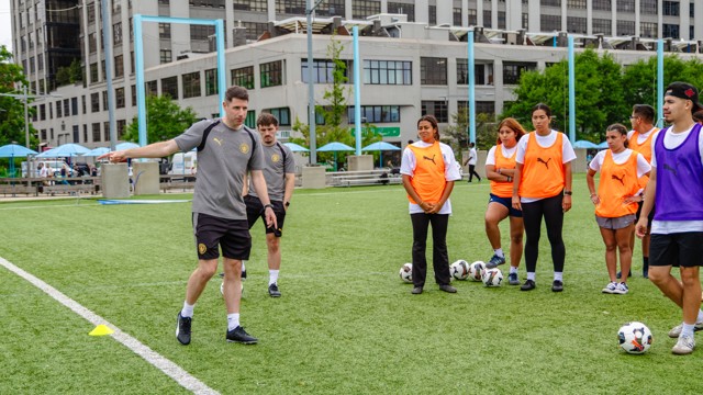 A soccer coach demonstrates techniques on a field to young players wearing orange and purple training bibs.