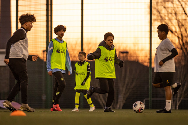 A group of young players practicing football on a pitch at sunset, wearing jerseys with the Premier League Kicks logo.