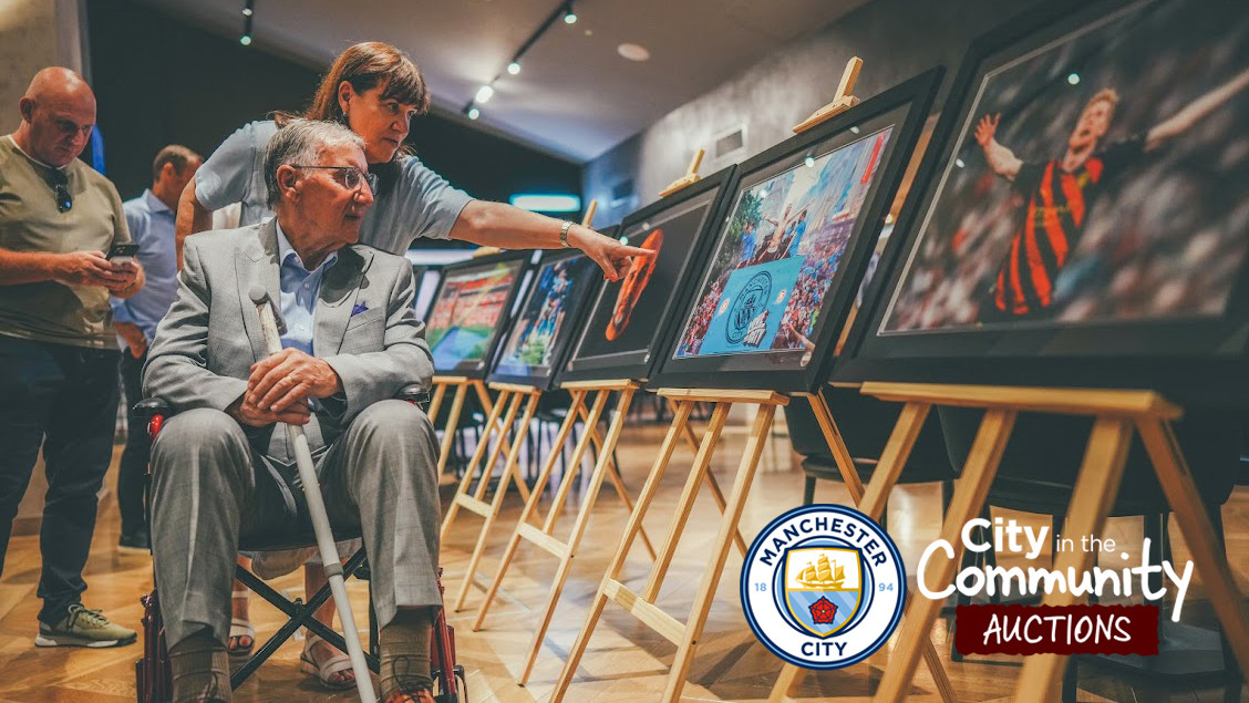 People viewing framed photographs at a Manchester City event with 'City in the Community Auctions' logo displayed.